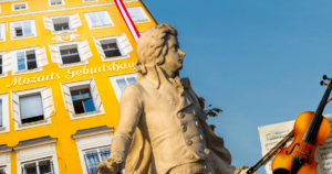 Statue von Mozart vor blauem Himmel, im Hintergrund Geburtshaus Mozarts und eine Geige mit Notenblättern